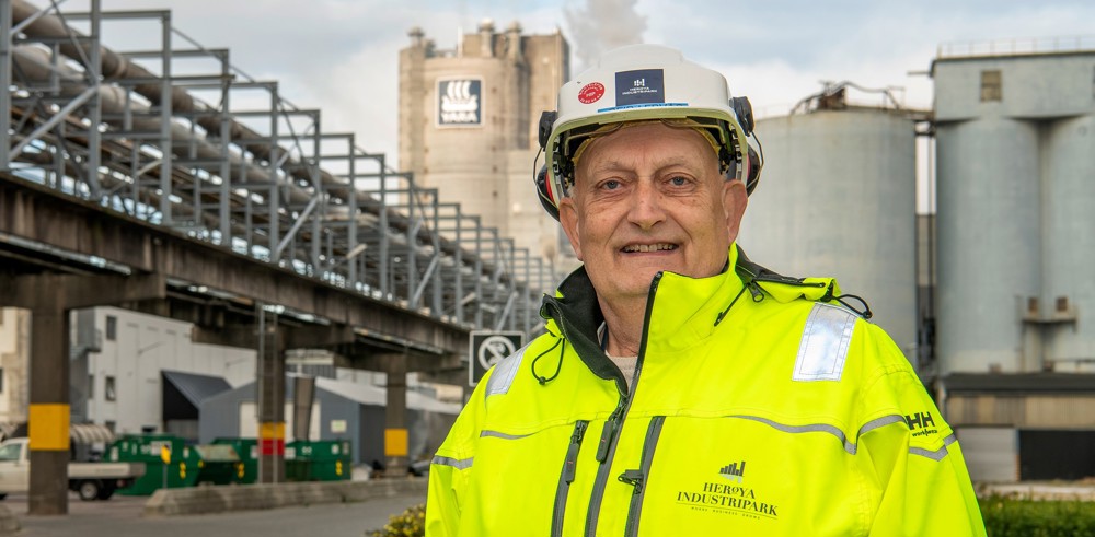 mann i gul jakke og hvit hjelm, poserer, industripark, rørbro og fabrikkbygg i bakgrunnen